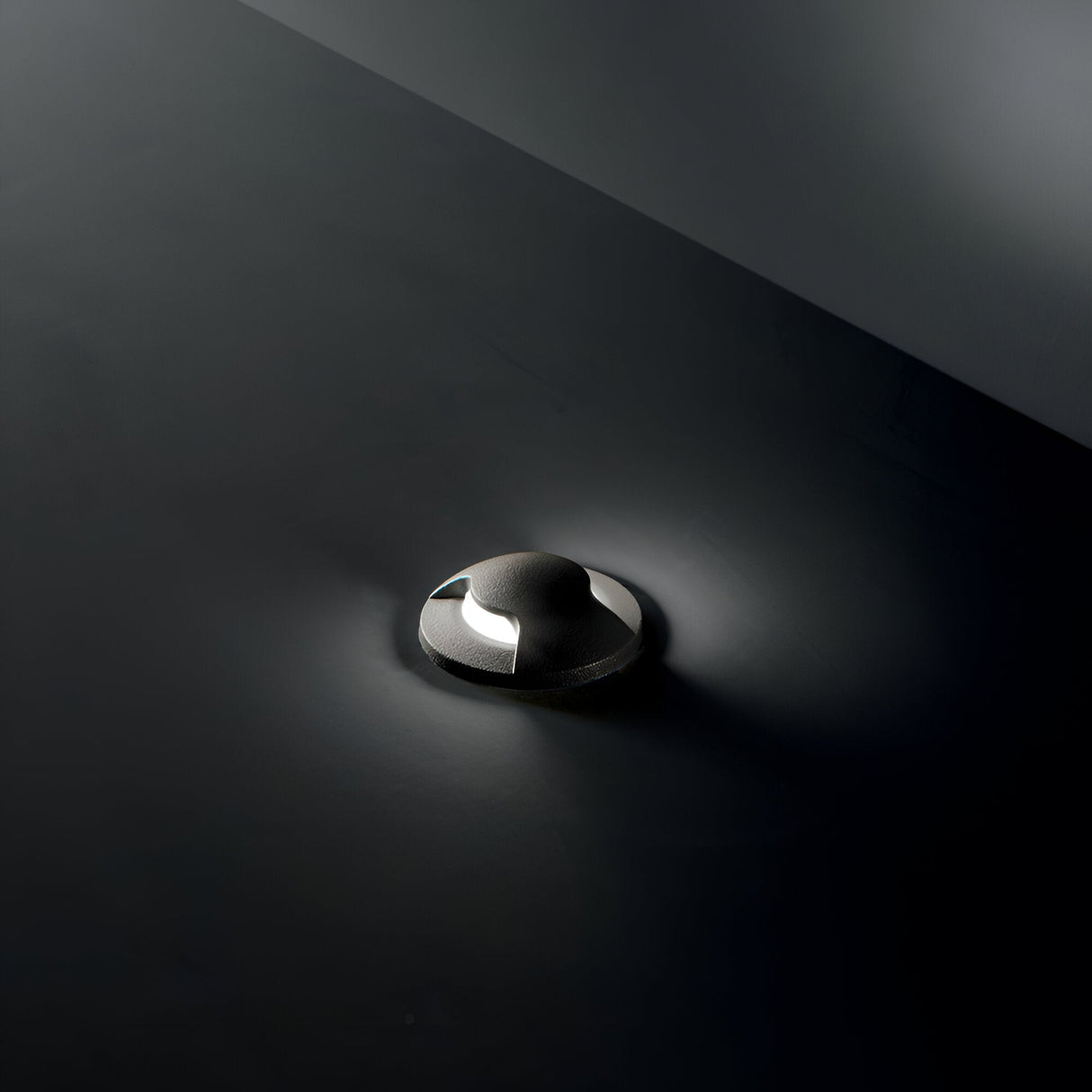 Against a dark background, the Solarium Outdoor Drive Over Recessed Light Two-Sided 3000K in Black casts a soft, ambient glow with its dimly lit brilliance. Shadows accentuate its sleek, metallic surface and elegant design. This luminaire mirrors the high-performance resin typical of outdoor fixtures, underscoring both durability and style.