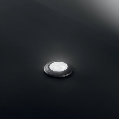 The Solarium Outdoor Drive Over Recessed Light 3000K in black, featuring a small, round design set against a dark background, emits a soft white glow. With an IP67 rating for outdoor use, this light offers subtle illumination. Its minimalist and modern design is crafted from high-performance resin, ensuring both durability and style.