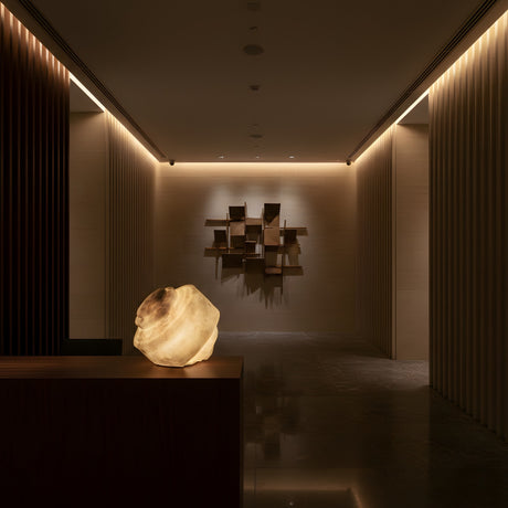 A modern, dimly lit hallway features the Selmora LED Fluid Silhouette Alabaster Table Lamp - Soft White on a dark table in the foreground, with geometric wood art at the end and warm recessed lights among vertical wood wall panels for a calm ambiance.