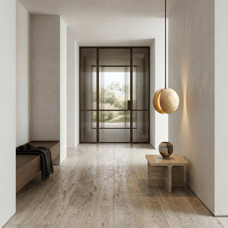 Minimalist hallway with stone floor, wooden bench draped in a dark blanket, small table with round sculpture, and the Zanvera 1 Light Split Orb Travertine Single Pendant Light filling the space with warm light and views of lush greenery outside.