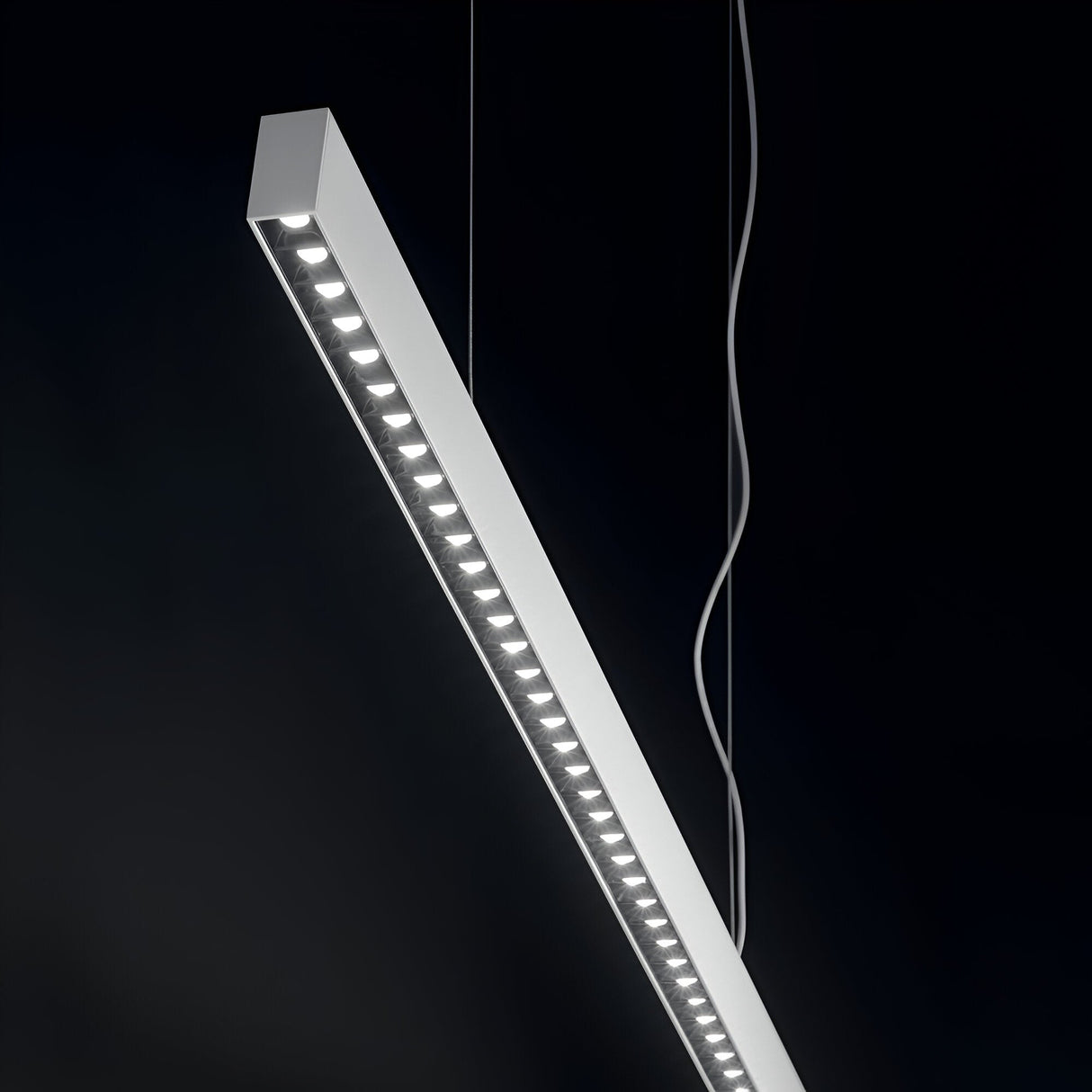 The Celine LED Linear Suspension 30W 3000K in white, with its modern and sleek design, epitomizes energy-efficient lighting as it hangs against a dark background. Suspended by two cables, the illuminated bulbs are evenly spaced along its length, demonstrating a perfect blend of sophistication in modern lighting.