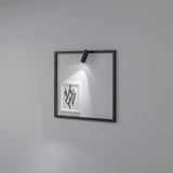 A minimalist frame featuring a small geometric pattern is mounted on a gray wall, subtly illuminated by the Glimmer LED Square Wall Light 5W 3000K in black. The LED light enhances the artwork with gentle shadows, and its square shape and simple, modern design make it an ideal contemporary lighting solution for any space.