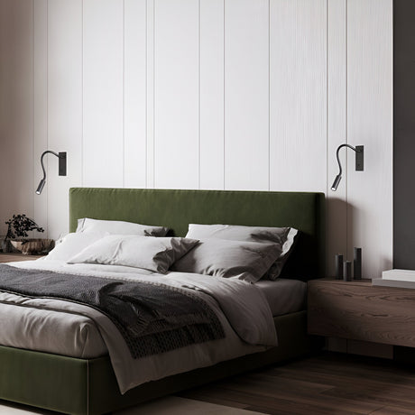 A modern bedroom showcases a green upholstered bed, gray bedding, and wood flooring. Enhanced by the Celeste LED Recessed Wall Light 3W 3000K in black above the bed on a white paneled wall, along with a wooden nightstand featuring minimal decor, it captures the essence of contemporary and minimalist style.