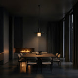 A modern dining room with a rectangular table, eight chairs, dark walls, tall curtains, a warm fireplace, and the Norevia 1 Light Cube Wide Alabaster Single Pendant Light in Soft White & Matte Black creates a moody, minimalist vibe.