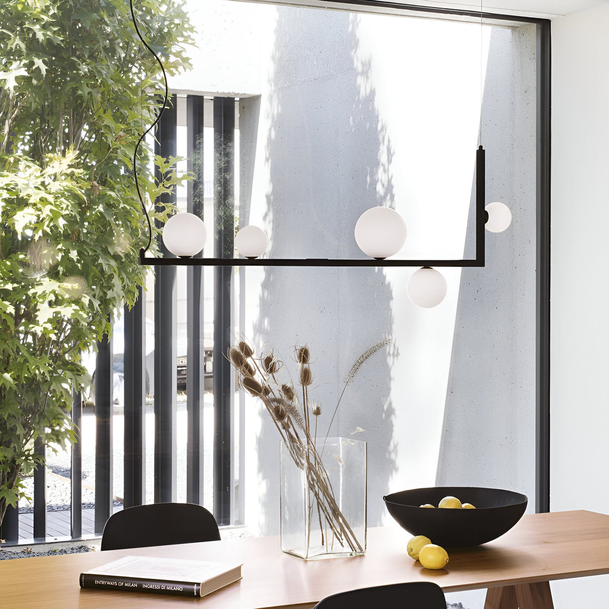 A modern dining area with a wooden table features a tall glass vase filled with dried plants and a black bowl of lemons. Above, the Ethereal 5 Light Suspension in black showcases sleek industrial design, hanging near large windows that provide a view of the greenery outside.