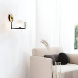 A minimalist room showcasing a light beige armchair, a black cylindrical pillow, and a glass vase with ornamental grass. The Ethereal Wall Light - White, with its sleek round bulb, complements the white curtains that gently filter soft natural light inside.