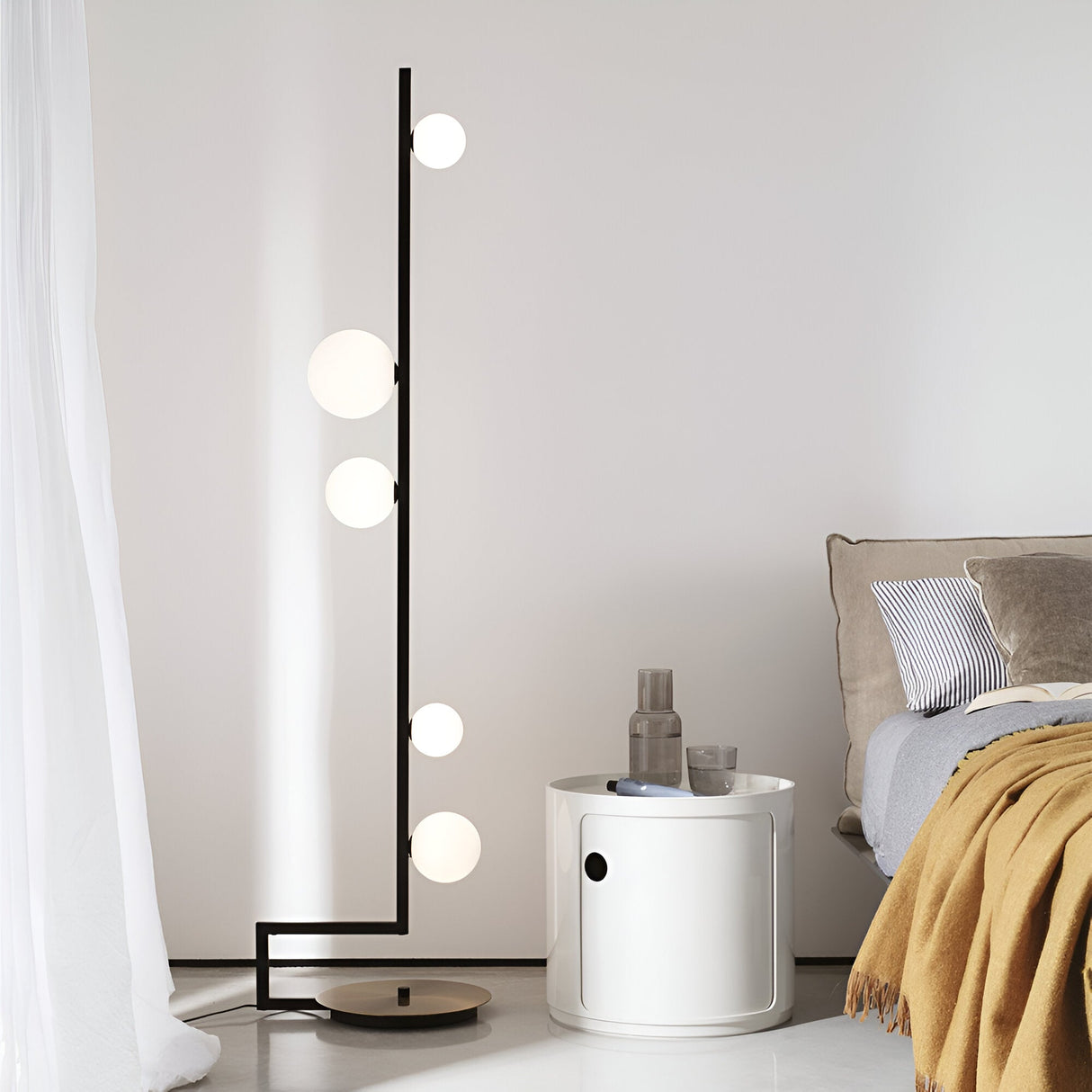 In a modern bedroom setting, an Ethereal Floor Lamp - Black/White features five globe lights on a vertical black pole. Nearby, a round white nightstand holds two glasses and a carafe. The sleek, modern design is completed with a beige bed decorated with decorative pillows and accented by a mustard throw blanket.