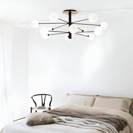 In a minimalistic bedroom, the Veloxis 12 Light Ceiling Light in black and white serves as a modern chandelier with its matte black frame and multiple spherical lights. This fixture includes a dimmable feature for customizable ambiance. The room also features a bed adorned with beige and white bedding, a black chair in the corner, and white curtains illuminated gently by natural light.
