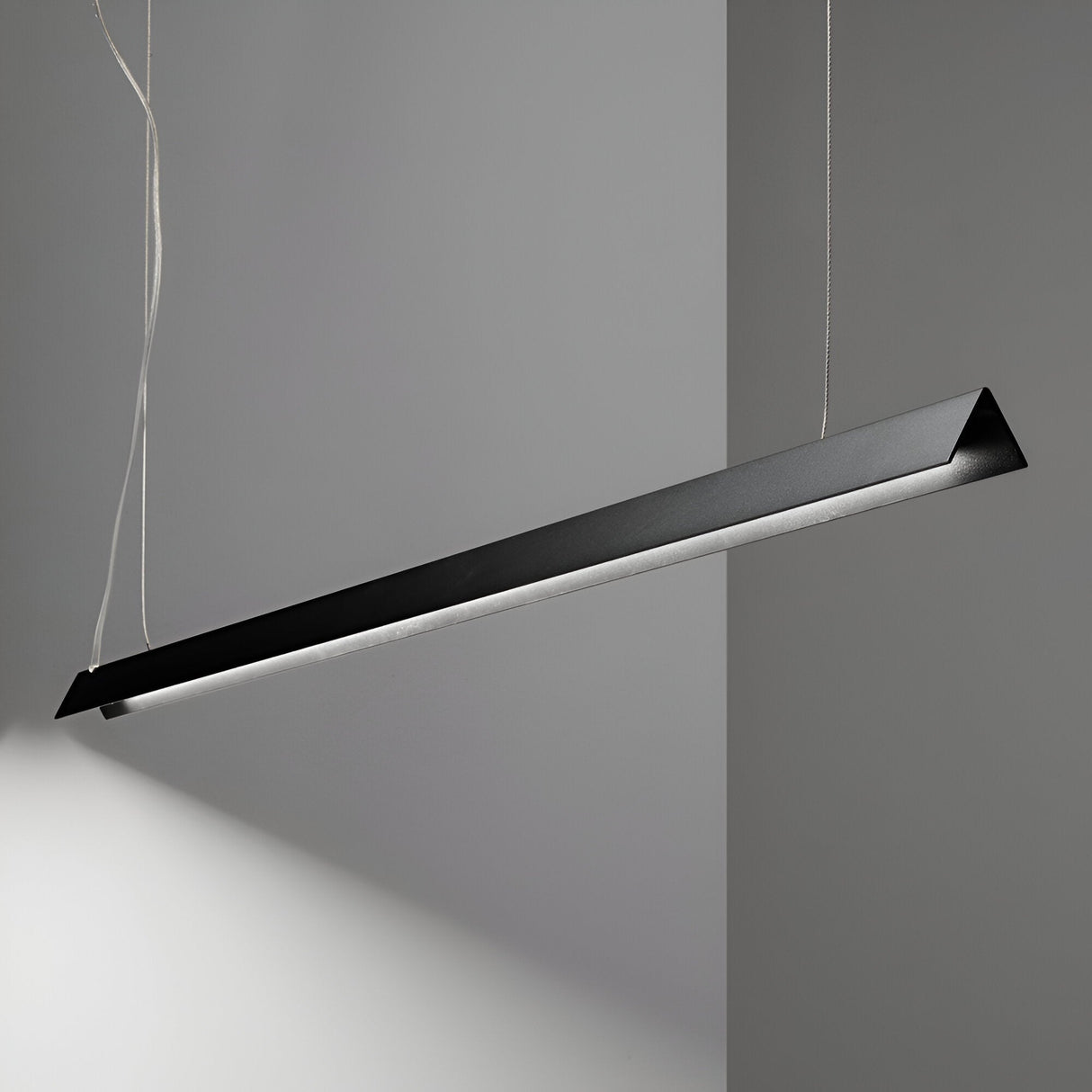 Suspended from the ceiling, the sleek Flicker LED Linear Suspension 20W 3000K - Black illuminates a gray wall with its energy-efficient lighting. This modern lighting fixture features a minimalist design with a long, narrow shape and clean lines, held by thin wires.