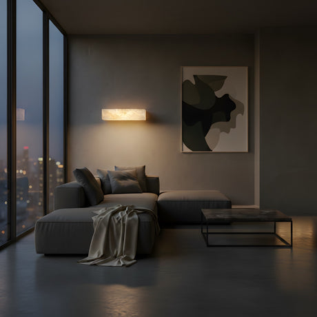 A modern, minimalist living room at dusk with floor-to-ceiling windows, a gray sectional, marble coffee table, wall art, and the Caelith LED Small Alabaster Shelf With Light – Soft White. A rectangular wall light and city skyline complete the tranquil space.