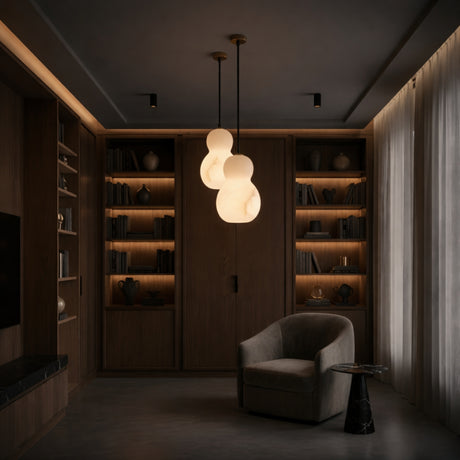 A cozy, modern reading nook features a curved armchair, round side table, and the Orlune LED Sculpted Pear Alabaster Single Pendant Light - Soft White. Built-in wooden shelves with books and decor are softly illuminated by warm lighting.