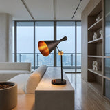 A Funk Table Lamp - Black sits on a concrete side table in a chic living room with large windows, city views, a white sofa, wooden shelves, and abstract white sculptures.