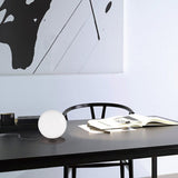 Libra Globe Glass Table Lamp with white spherical glass diffuser on a black metal base, shown on a dark desk beside a chair and open book.