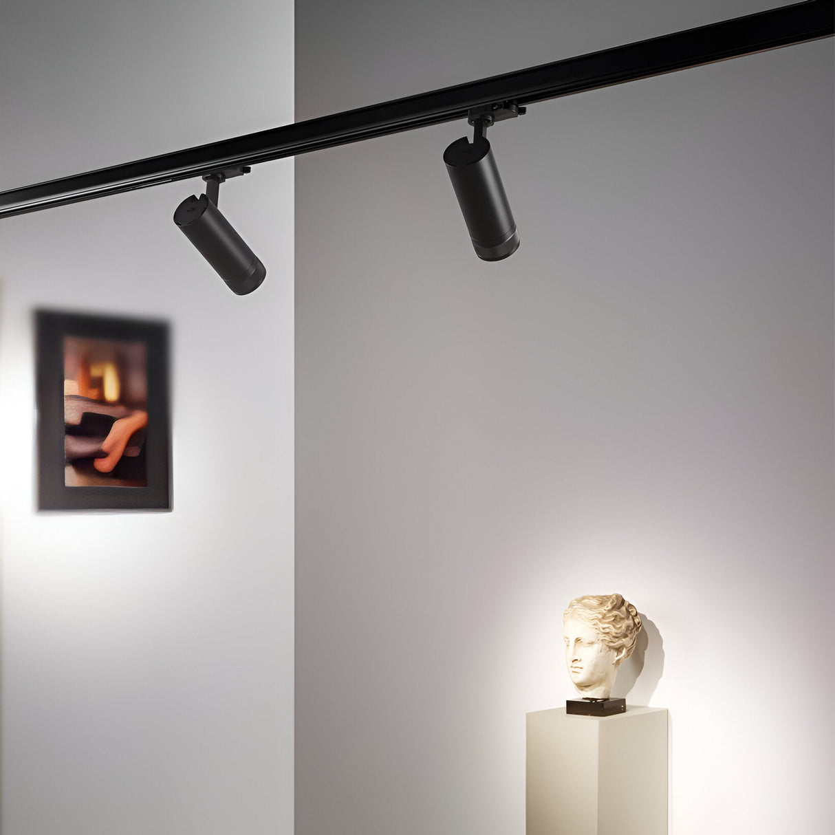 In a modern art gallery corner, a small bust rests on a pedestal under the glow of an adjustable lighting setup. Two Solis Three Phase LED Track Spotlights 15W 3000K in black project a focused light. In the background, a blurred framed artwork partially hangs visible on the wall.