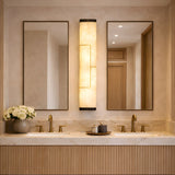 Elegant bathroom with double sinks, two rectangular mirrors, the Eryndor LED Alabaster Wall Light in matte black & soft white, gold fixtures, and white flowers on a marble countertop. Warm, neutral tones complete the space.