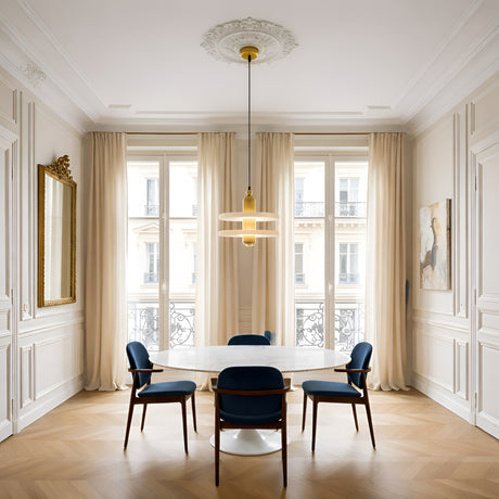Elegant dining room featuring a round white marble table, four blue chairs, light wood floors, tall windows with sheer curtains, a gold-framed mirror, wall art, and the Nyxara LED Small Double Tiered Alabaster Single Pendant Light in soft white & gold.