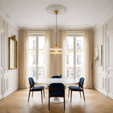 Elegant dining room with a round white marble table, four blue upholstered chairs, large windows with sheer beige curtains, a gold-framed mirror, wall art on the right, and the Nyxara LED Large Double Tiered Alabaster Pendant overhead.