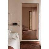 Lucka Bronze Tinted Full Length Mirror 200cm in a black metal frame, leaning against a bedroom wall and reflecting a warm-toned interior with curtains and herringbone wood flooring.