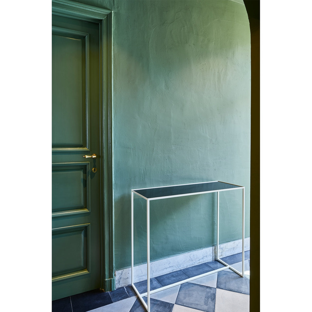 Mesa White console table with slim white metal frame and reversible mirrored glass top in dark grey, shown against a green wall in a hallway setting.