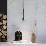 In a minimalist interior space with a concrete wall, the Solenia LED Light Pendant 7W 3000K in black provides an energy-efficient modern lighting solution. Nearby, a stack of firewood rests next to "Sottsass" on a shelf, accompanied by a minimalist sculpture and a glass vase with dried flowers.