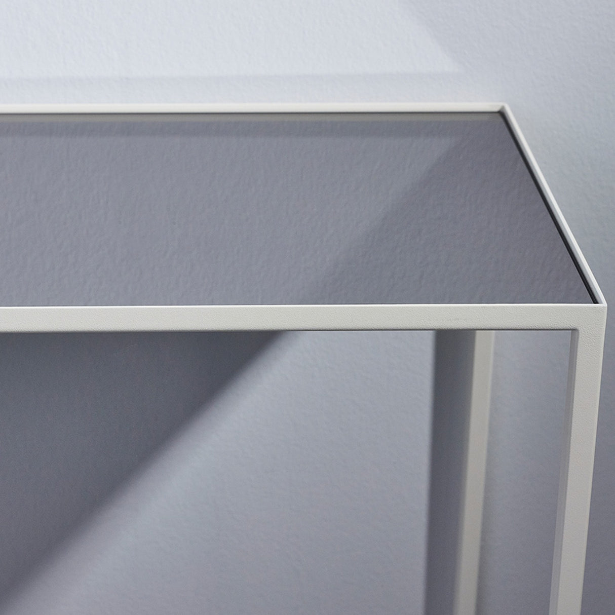 Close-up of the Mesa White console table's corner, showing the slim white metal frame and grey-tinted mirrored glass top against a light grey background.