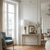 Elegant Parisian-style living room with a vintage blue armchair, tall window with beige curtains, ornate white walls, and a console table featuring the Valmora 1 Light Monolith Marble Table Lamp in brass & white, gold-framed mirror, and greenery-filled vase.
