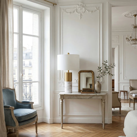 Elegant Parisian-style living room with a vintage blue armchair, tall window with beige curtains, ornate white walls, and a console table featuring the Valmora 1 Light Monolith Marble Table Lamp in brass & white, gold-framed mirror, and greenery-filled vase.