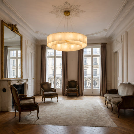 Elegant living room with high ceilings and large windows. An Arvello 8 Light Layered Panel Alabaster Chandelier (Soft White & Brass) glows above vintage armchairs, a sofa, beige rug, and marble fireplace topped by a gold-framed mirror.
