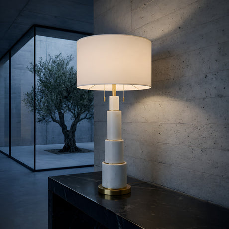 The Eloria 1 Light Stacked Alabaster Table Lamp in Brass & Soft White sits on a dark marble surface in a minimalist room. A large window reveals an olive tree outside against concrete walls.