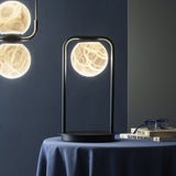 Halvessa LED Alabaster Dimmable Table Lamp in black, featuring a glowing alabaster globe held within a slim arched iron frame on a round base, shown on a bedside table in a dark-toned room.