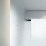 Close-up of the B.Ambi Plus 70cm square backlit LED demister mirror's edge, showing the frameless glass corner mounted on a white bathroom wall beside a hanging white towel.