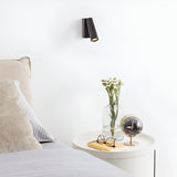 A modern bedroom scene features a bedside table adorned with a book, glasses, a vase of flowers, and a decorative clock. Nearby, a bed with beige and striped pillows complements the aesthetic. Mounted against the white wall above the table is the Radiant LED Single Spotlight 7W 3000K in Black, providing adjustable lighting.