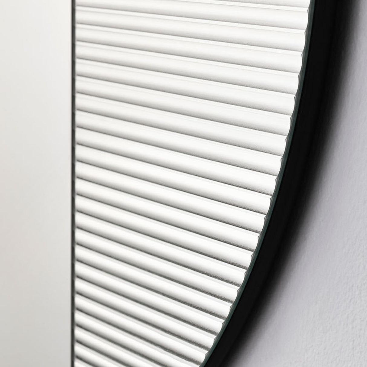 Close-up of the Cord Flutes round wall mirror, 58cm, showing horizontally ribbed reeded glass with a thin black frame edge against a plain white wall.