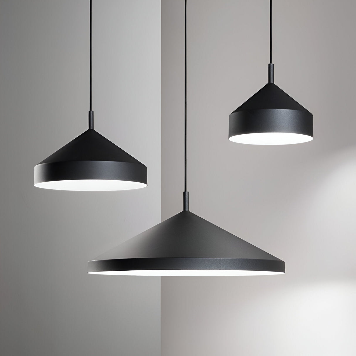 A trio of Elysian 1 Light Pendants in black, each with a sleek conical design and measuring 21cm, are suspended at varying heights against a minimalist white backdrop. These illuminated lamps cast gentle light and shadows on the wall, making them an ideal addition to minimalist interiors.