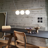 In a contemporary dining space, a sleek black table is paired with two wicker chairs against a concrete wall. Overhead, the Zephira 3 Light Linear Suspension in brass gently lights up the area with dimmable G9 bulbs. Wooden bowls filled with plums introduce warmth, while a tall mirror leans elegantly against the wall.