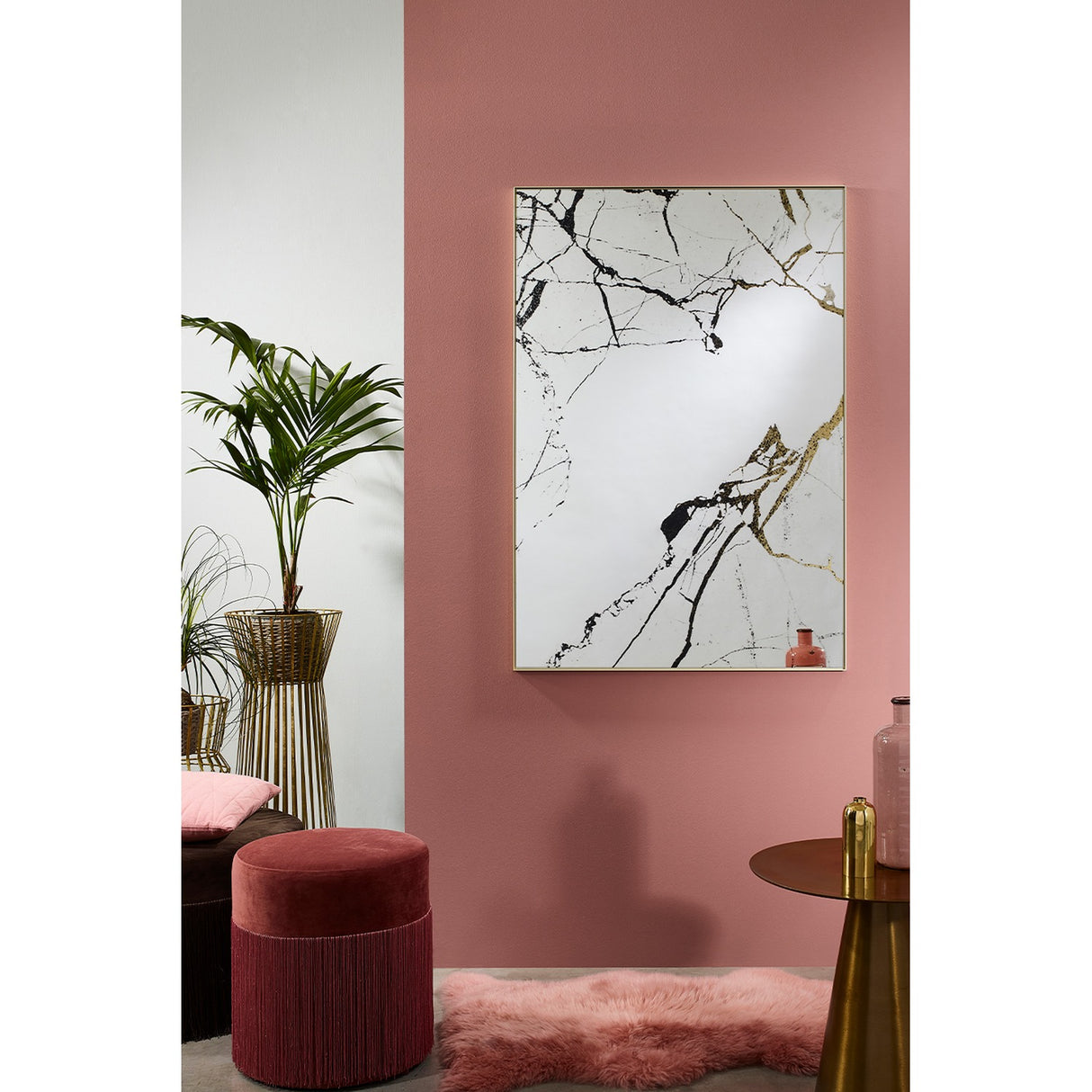 Rectangular wall mirror with gold frame, marble-effect veining printed on the glass, hung on a pink wall in a living room styled with a plant and red stool.
