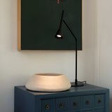 The Solenia LED Table Lamp 6.5W 3000K - Black, an energy-efficient fixture with a curved shade, casts light on a small wooden cabinet featuring multiple drawers. Atop the cabinet sits a round, beige ceramic bowl, while a dark green and black abstract painting adorns the wall behind.