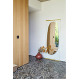 Obu Organic Full Length Leaning Mirror 178cm in gold, with an organic rock-shaped mirrored area set within a clear glass rectangle, leaning against a wall in a hallway setting.