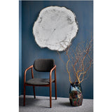Arbo Dark irregular frameless wall mirror with tree-ring print design, mounted on a blue wall above a wooden armchair in a living room setting.