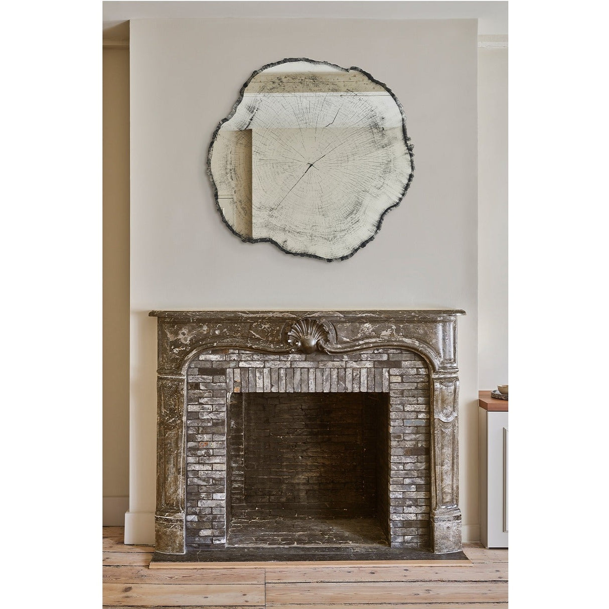 Arbo Dark irregular frameless wall mirror with tree-ring printed glass face, hung above a stone fireplace with shell carving in a living room setting.