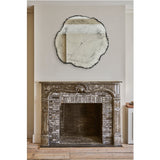 Arbo Dark irregular frameless wall mirror with tree-ring printed glass face, hung above a stone fireplace with shell carving in a living room setting.