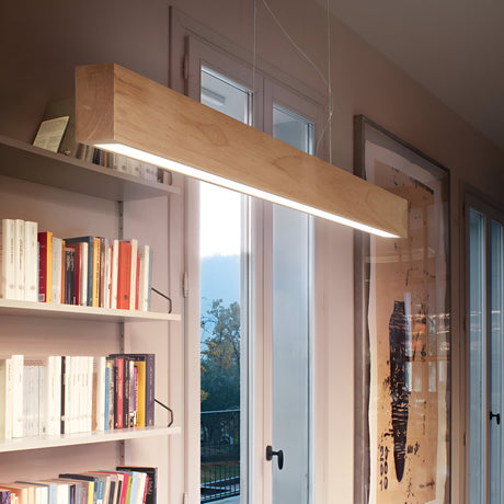 A modern interior showcases the Erin LED Wood Linear Suspension 37W 3000K in Light Brown hanging from the ceiling. The room features a large window with views of trees, a bookshelf filled with books on the left, and framed artwork leaning against the wall, all enhanced by energy-efficient lighting.