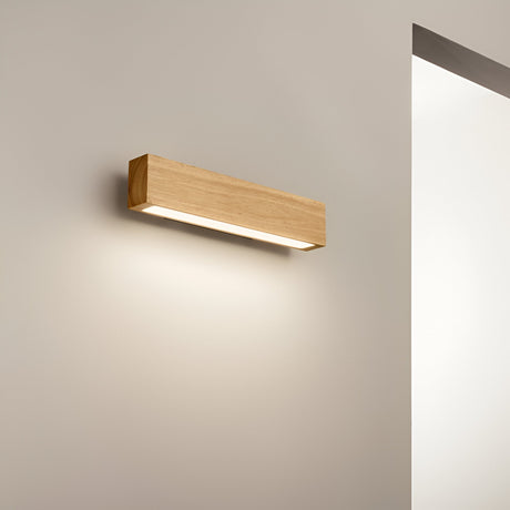 The Erin LED Wood Wall Light 13W 3000K in light brown, measuring 40cm, is affixed to a cream-colored wall, emitting a cozy radiance. Its natural wood design enhances the modern lighting style, with the backdrop featuring a vertical corner where the wall transitions into a bright white area.