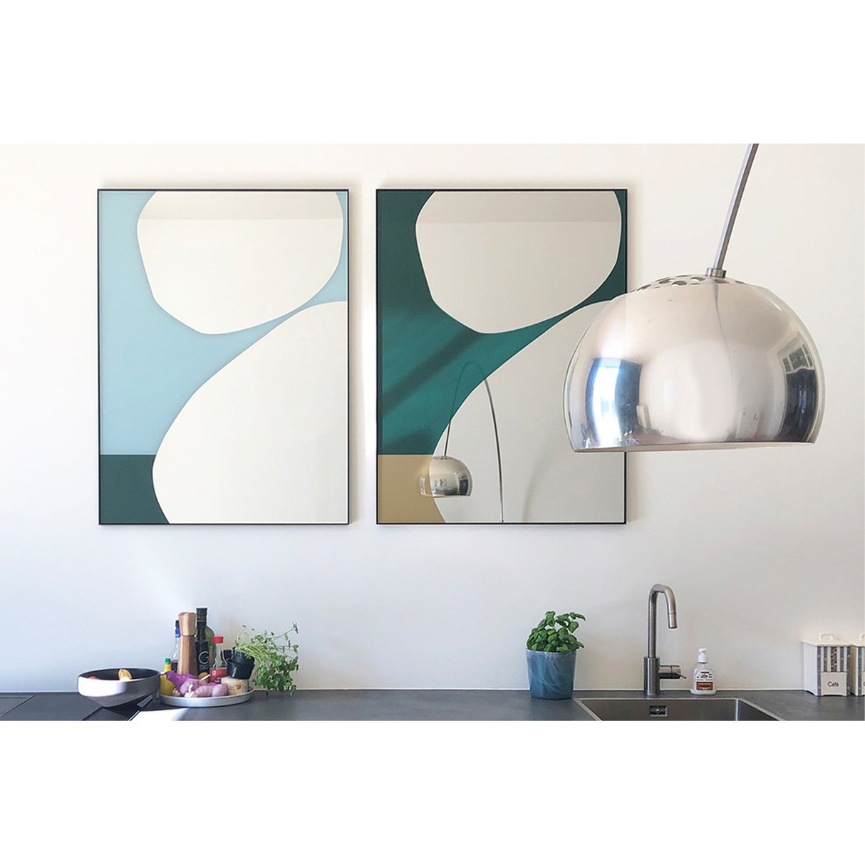 Mika Blue Printed Glass Wall Mirror 107cm in a slim black frame with teal and green curved shapes, shown beside a matching mirror on a kitchen wall.