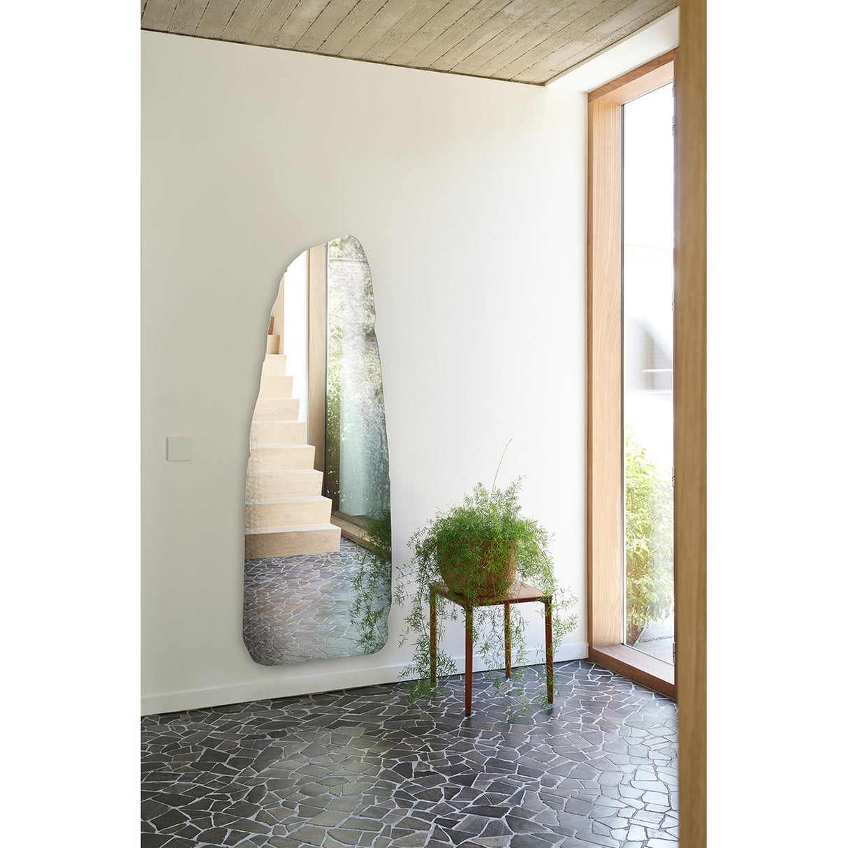 Rocca Standing Stone Printed Glass Wall Mirror 175cm, frameless irregular menhir-shaped mirror leaning against a hallway wall near a wooden staircase and potted plant.
