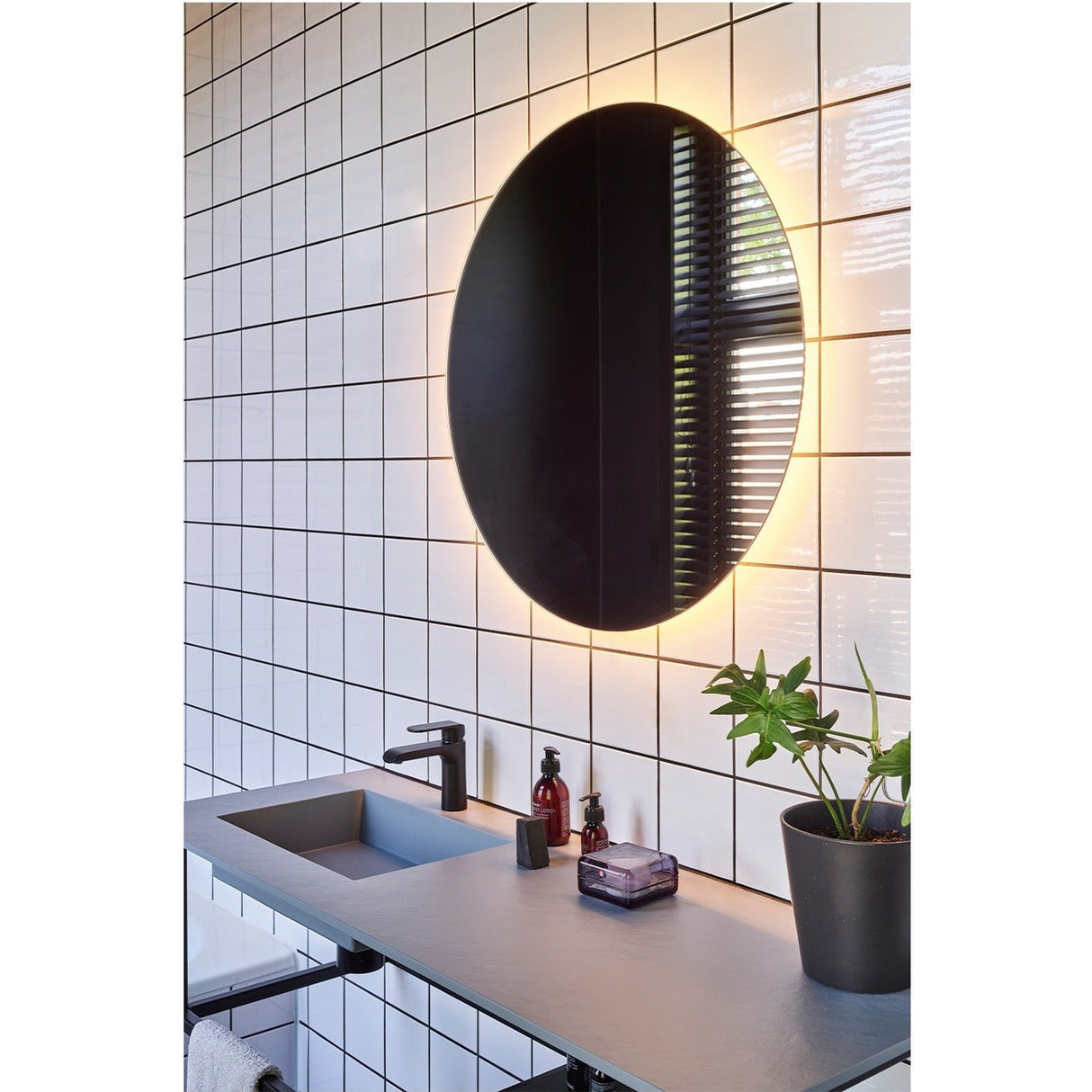 Brio Clear Round LED IP44 Bathroom Mirror, frameless round mirror with backlit LED glow, mounted above a bathroom vanity with black tap and tiled wall.