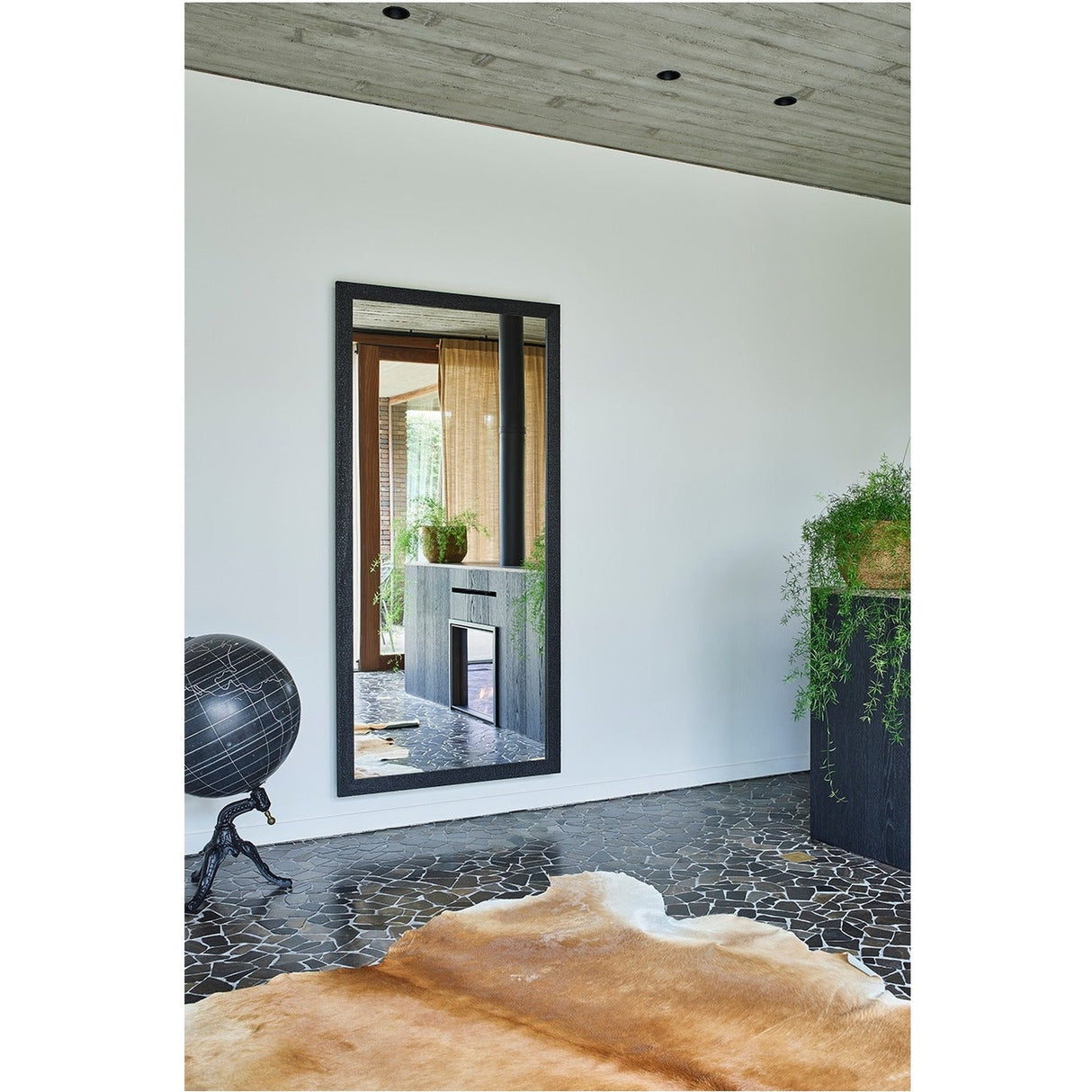 Kyo Wooden Full Length Mirror in Black leans on a white wall in a living room, rectangular black wooden frame reflecting a wood-burning stove and interior view.