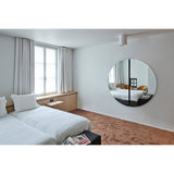 Luna Black Two Piece Round Wall Mirror 150cm above a bed in a bedroom, frameless glass disc split by a black lacquered central strip, reflecting the room.