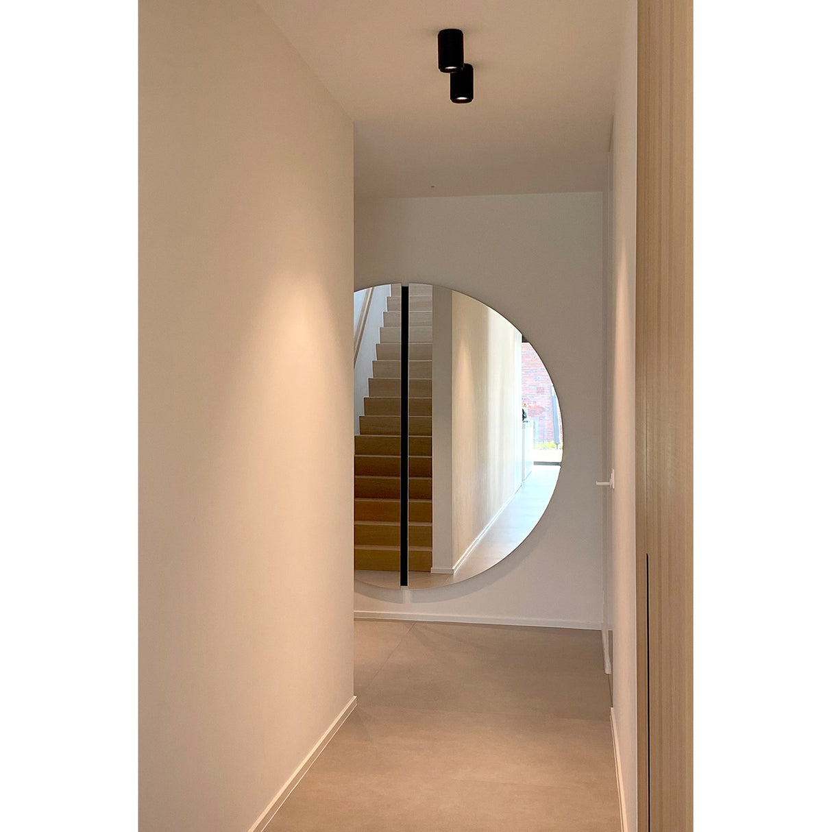Large round frameless wall mirror in a hallway, formed of two half-moon panels with a black lacquered glass strip marking the central join, reflecting a staircase.