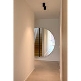 Large round frameless wall mirror in a hallway, formed of two half-moon panels with a black lacquered glass strip marking the central join, reflecting a staircase.
